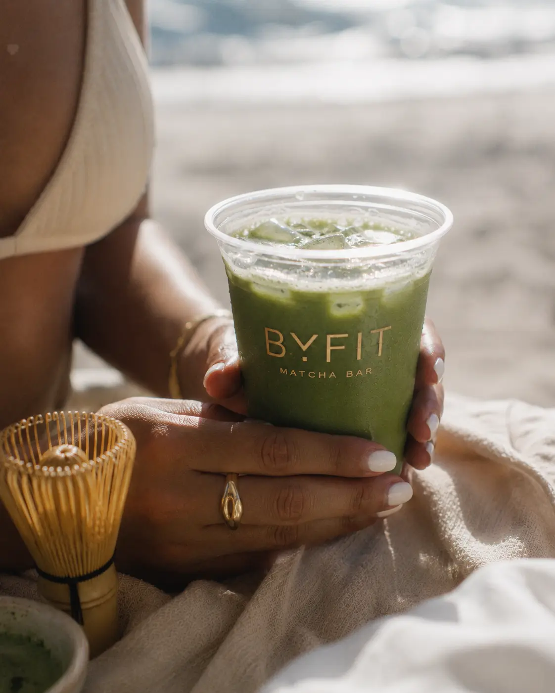 Matcha helado BYfit sostenido en la playa junto a un batidor de bambú